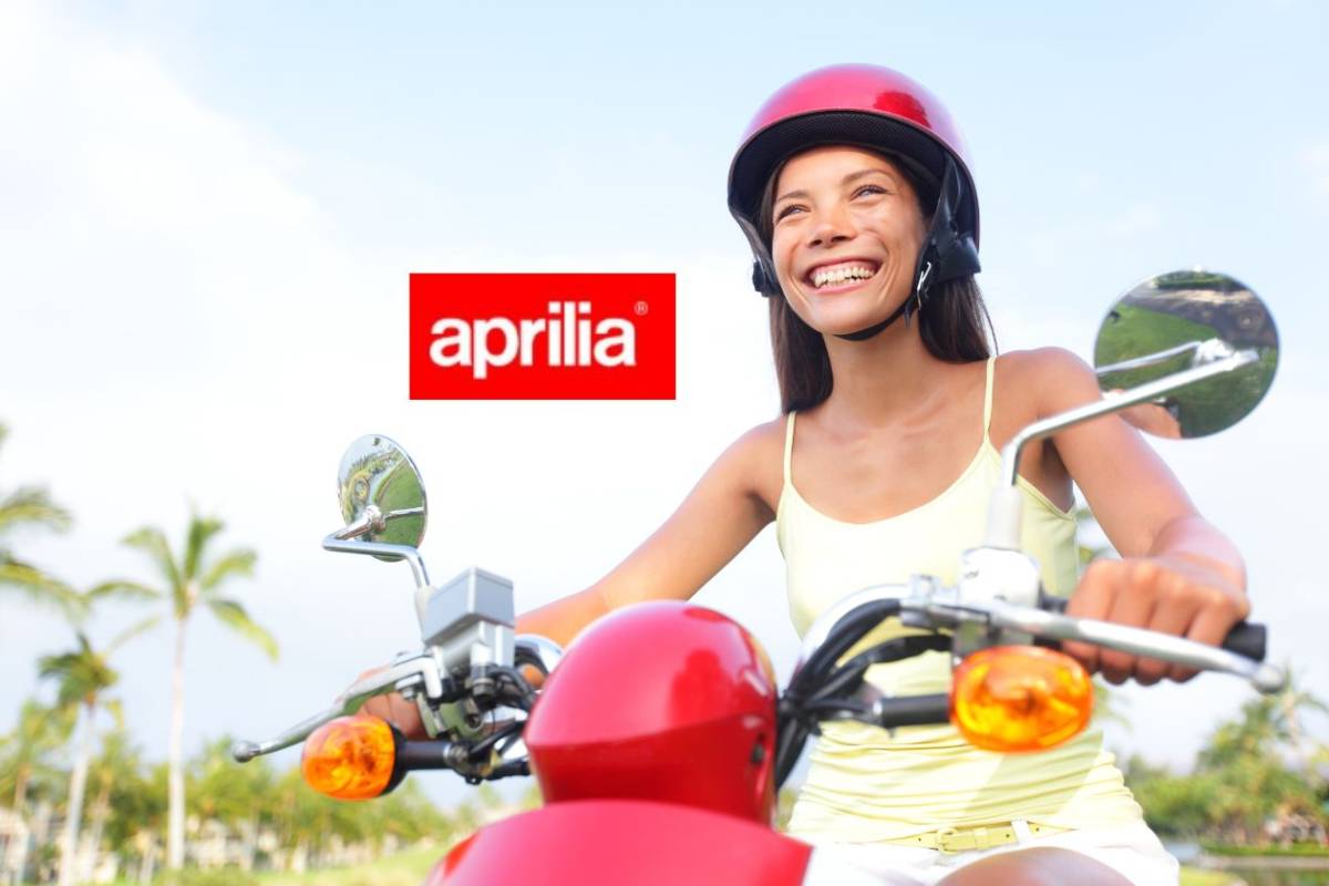 Una ragazza sorridente alla guida di uno scooter. A sinistra il marchio Aprilia