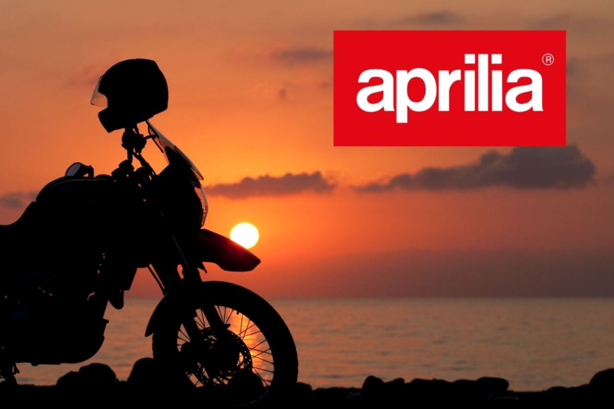 Una moto in ombra al tramonto sulla spiaggia con sopra un casco e a destra il marchio Aprilia